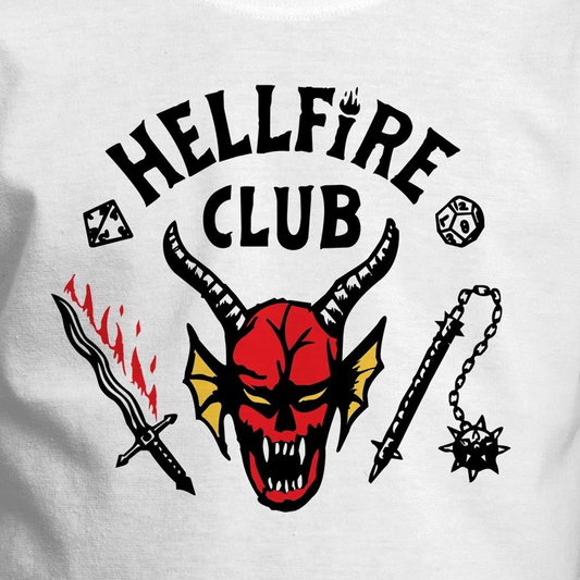 A close-up of a white t-shirt featuring a graphic print with the text 'HELL FIRE CLUB' in black arched lettering. Below the text is a red horned demon skull with yellow accents, flanked by a black sword with red flames, a black mace, and dice icons.
