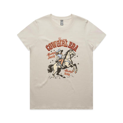 A white graphic t-shirt featuring a retro western illustration of a cowgirl riding a horse. The text reads 'In my COWGIRL ERA' in arched red letters, 'Always Sassy' in red script, and 'Always Wild' in red script, with the cowgirl wearing a cowboy hat and boots while riding a rearing horse.