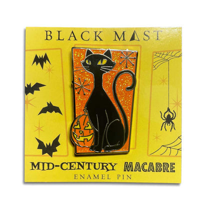 A Halloween enamel pin featuring a black cat sitting on a glowing orange pumpkin against an orange glittery background, mounted on a yellow retail card labeled 'BLACK MAEST' and 'MID-CENTURY MACABRE ENAMEL PIN'.