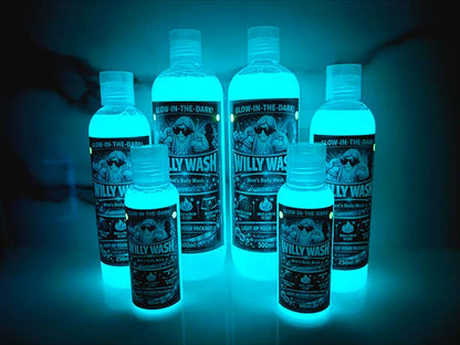 Six bottles of glow-in-the-dark body wash by Willy Wash arranged in a group, glowing with a bright blue-green light against a dark background.