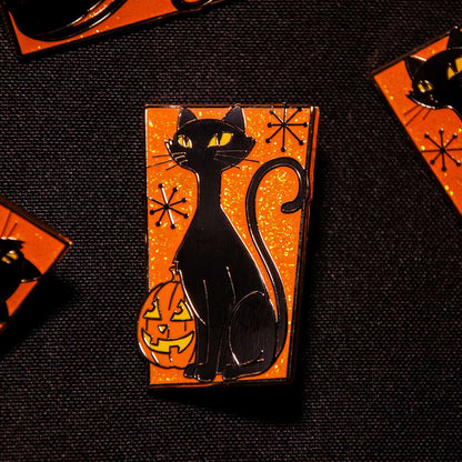 A rectangular enamel pin featuring a black cat sitting on an orange glittery background with a carved jack-o'-lantern and small black starburst motifs, displayed on a dark textured surface.