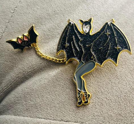 Pin with a design of a woman in a black dress with bat wings on a textured surface