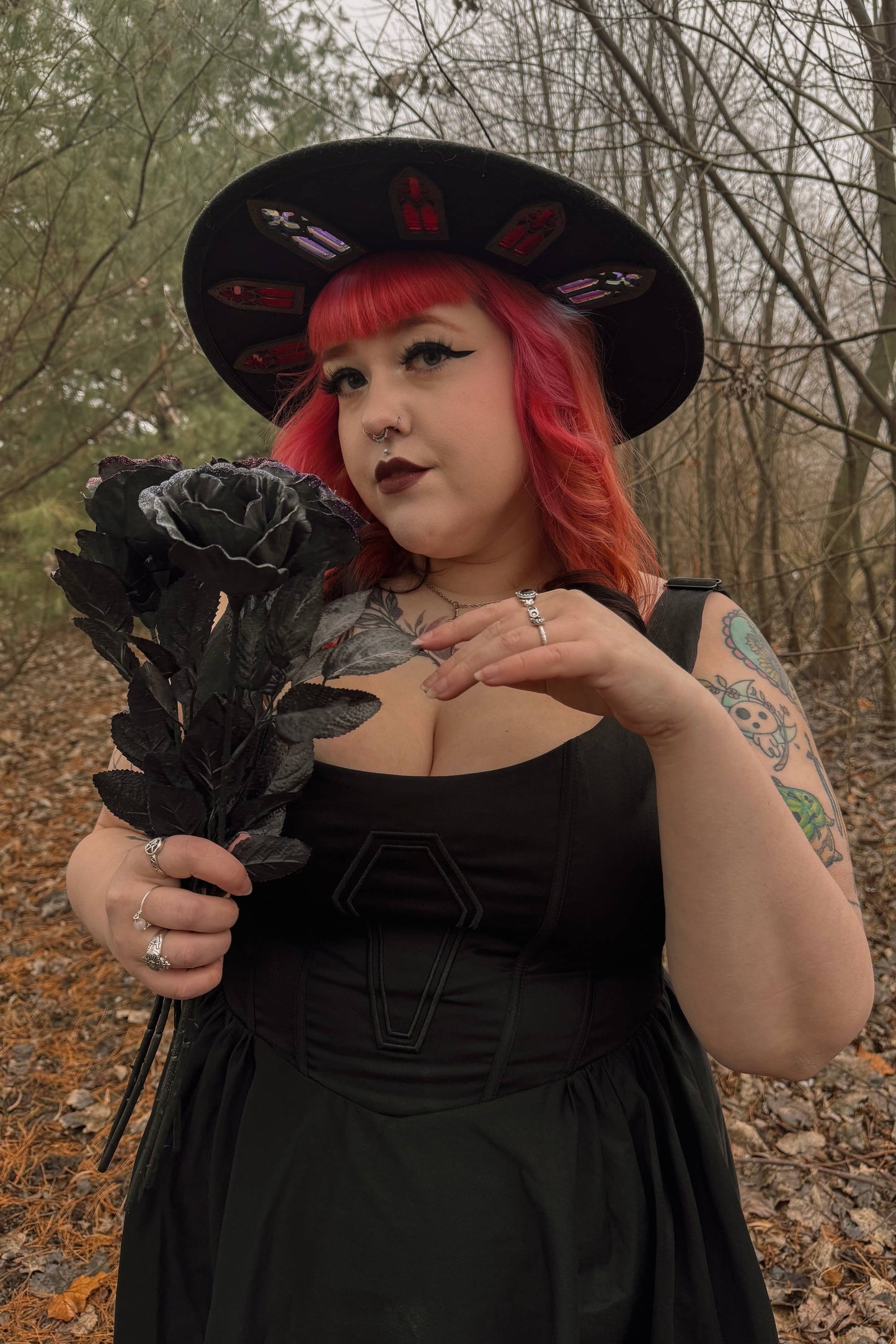 Person with red hair and black hat holding black roses in a forest setting