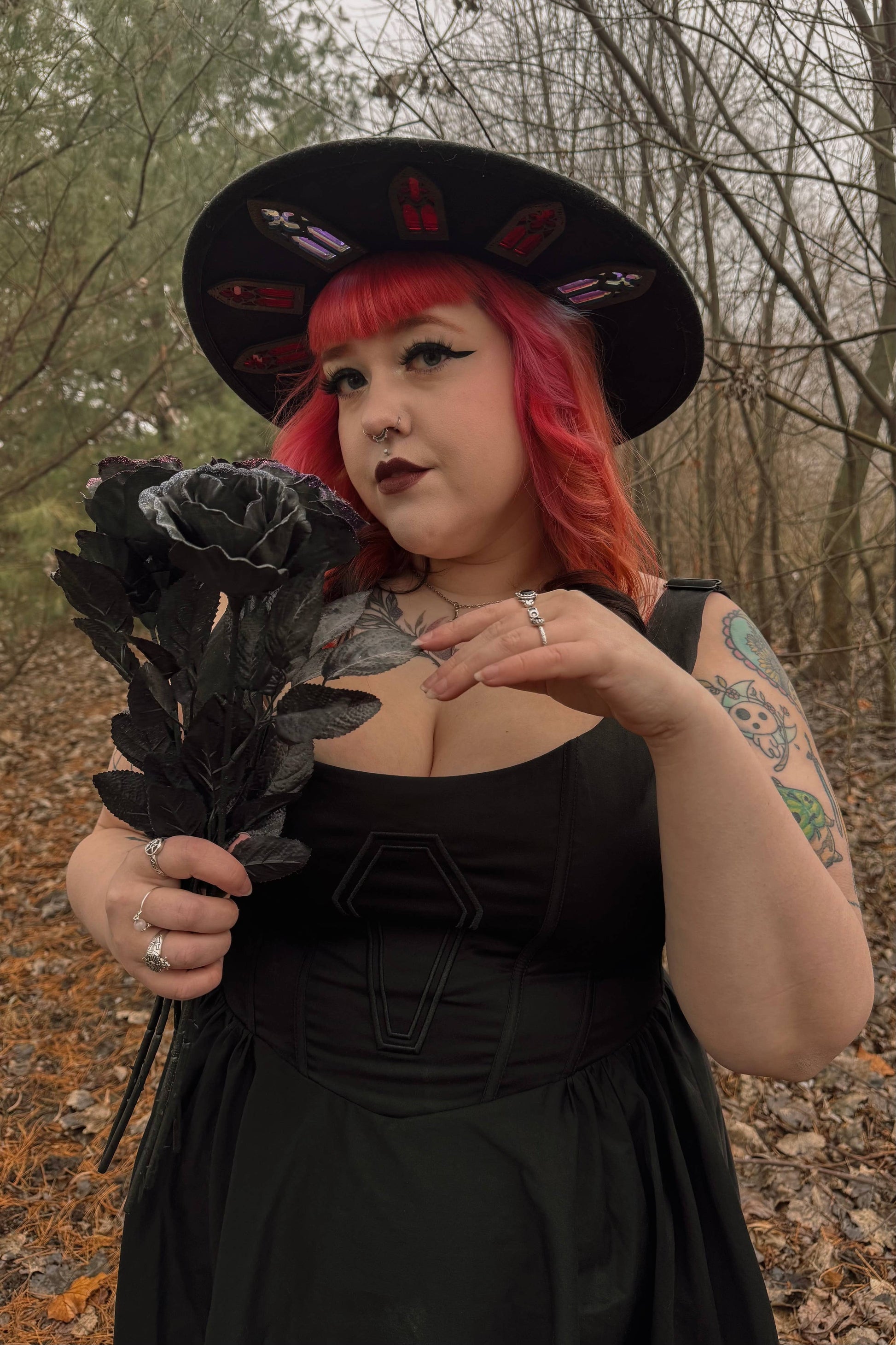 Person with red hair and black hat holding black roses in a forest setting