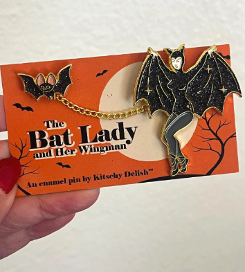 Enamel pin of a bat with a person's face on an orange card with Halloween design