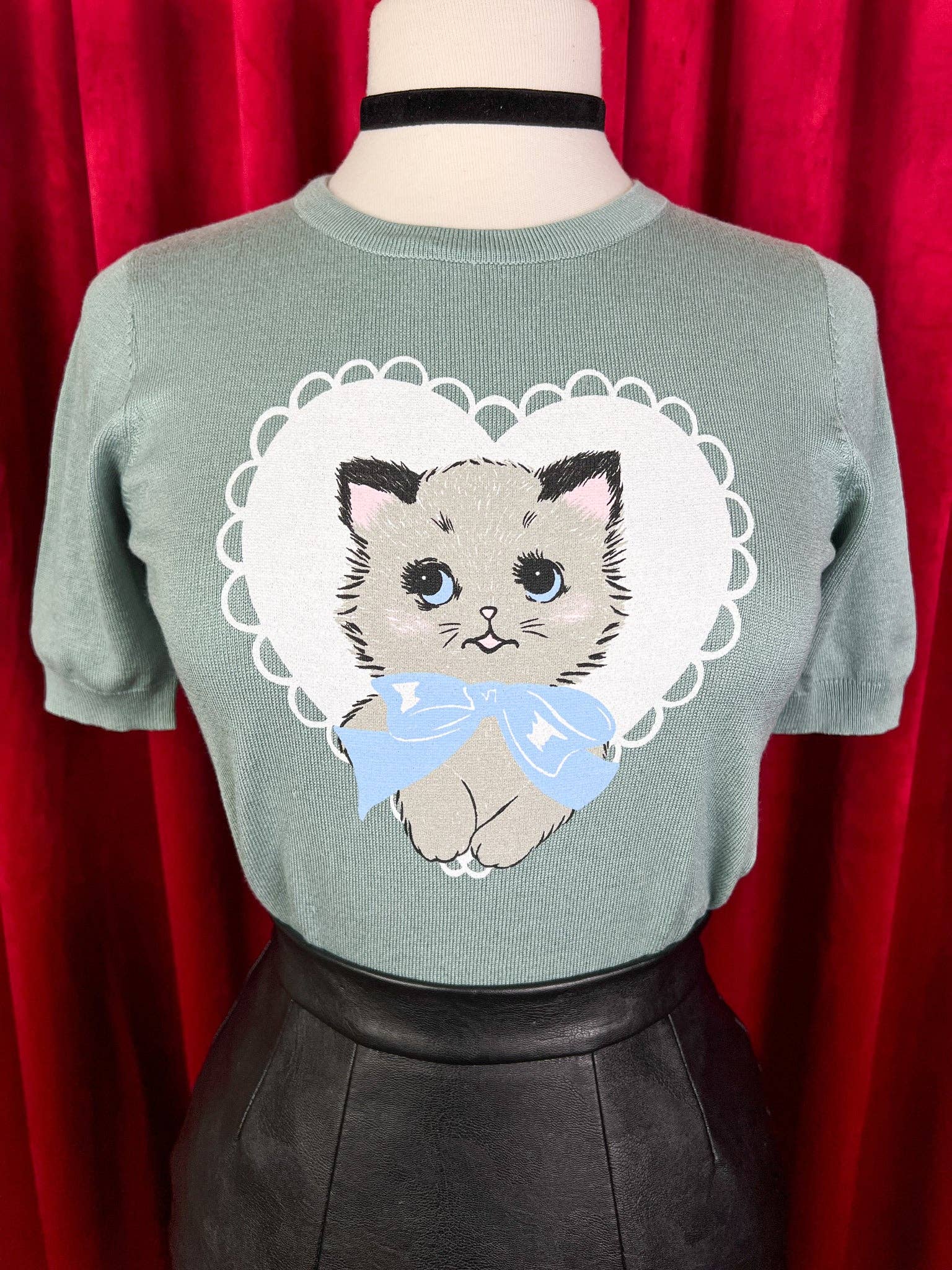 Green sweater with a cat design on a mannequin against a red curtain background