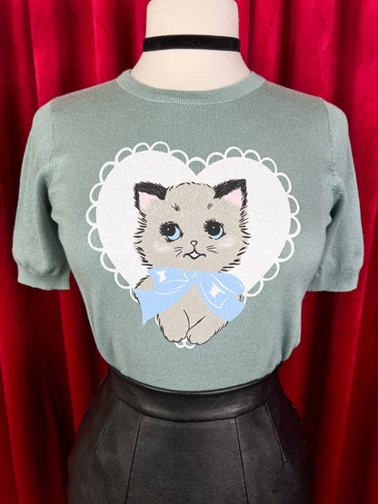 Green sweater with a cat design on a mannequin against a red curtain background