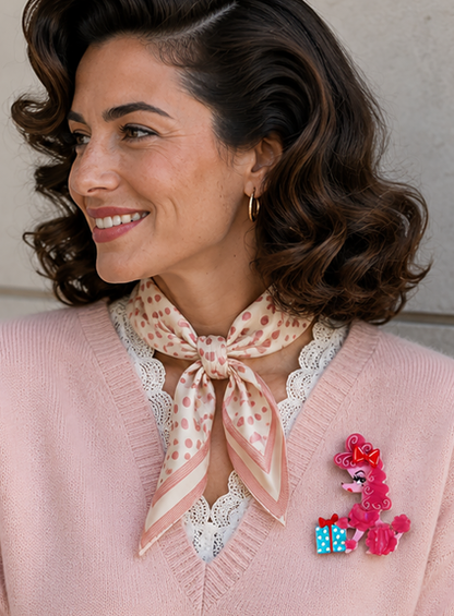 A woman with styled brown hair is shown in profile wearing a pink knit cardigan, a patterned neck scarf, and a bright pink poodle-shaped brooch with blue gift boxes.