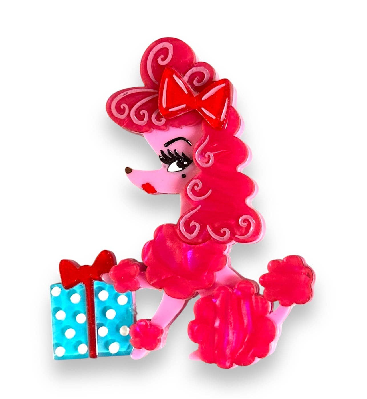 A pink poodle-shaped accessory, likely a brooch or charm, holding a small blue and white polka-dotted gift box with a red bow. The poodle features bright pink curly hair with white swirl patterns and a red bow, set against a plain white background.