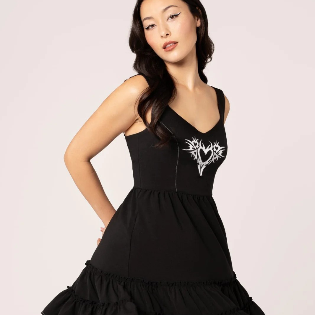 A woman modeling a black sleeveless dress with a V-neckline and a white graphic print on the chest, set against a solid light gray background.