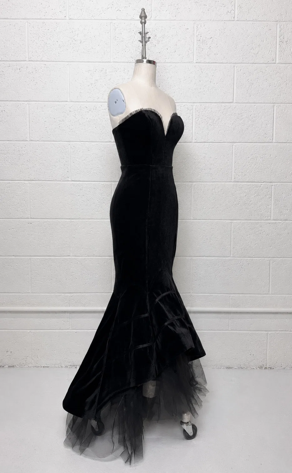A black strapless velvet gown with a sweetheart neckline and a flared tulle hem, displayed on a mannequin against a white painted brick wall.