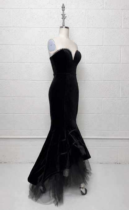 A black strapless velvet gown with a sweetheart neckline and a flared tulle hem, displayed on a mannequin against a white painted brick wall.