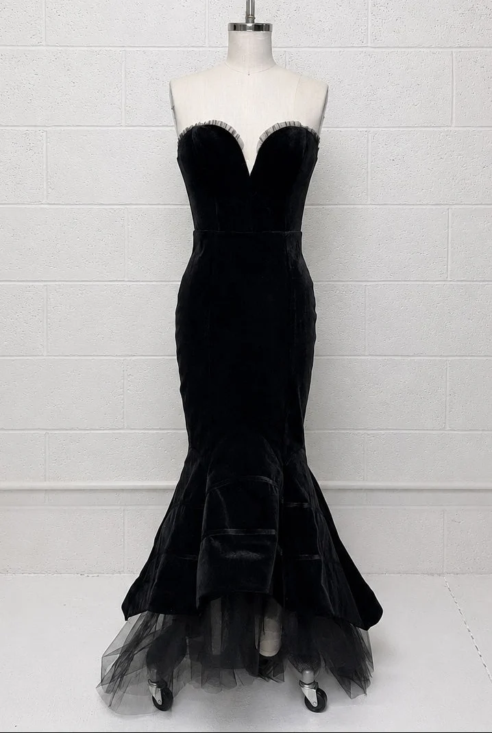 A black velvet strapless mermaid gown displayed on a white mannequin torso against a white painted brick wall. The dress features a sweetheart neckline with a thin silver beaded trim, a fitted bodice, and a flared tulle skirt.