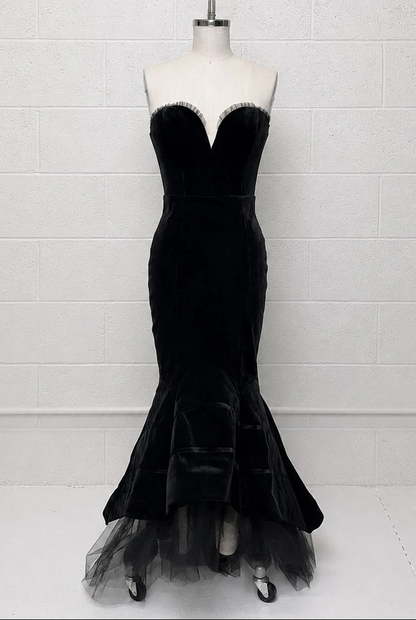 A black velvet strapless mermaid gown displayed on a white mannequin torso against a white painted brick wall. The dress features a sweetheart neckline with a thin silver beaded trim, a fitted bodice, and a flared tulle skirt.