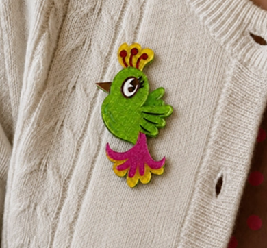 A colorful felt bird brooch with a green body, yellow and orange crown, and pink tail, pinned onto a cream-colored cable-knit sweater.