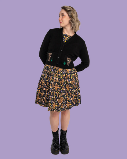 Person wearing a black cardigan with cow designs, a patterned skirt, and black boots on a purple background