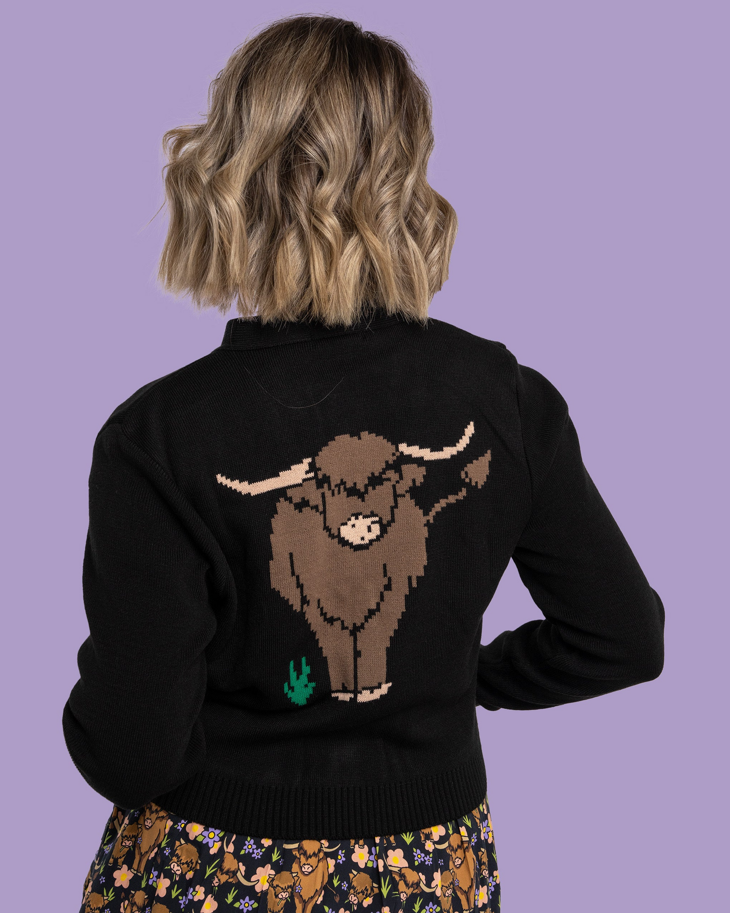 Person wearing a black sweater with a brown highland cow design on the back against a purple background
