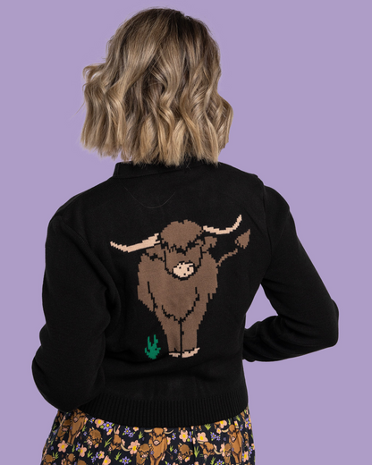 Person wearing a black sweater with a brown highland cow design on the back against a purple background