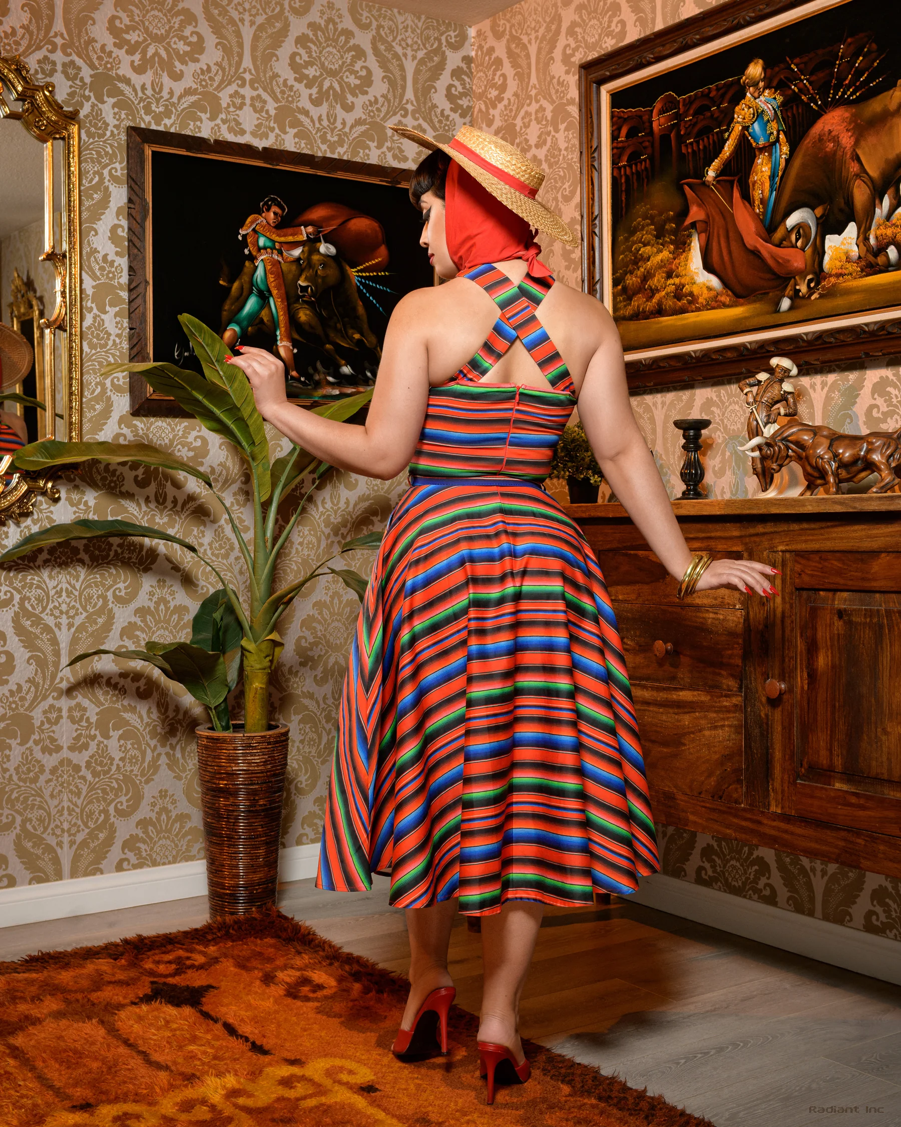 A woman stands indoors wearing a colorful striped halter dress with a flared skirt, a wide-brim straw hat, and a red headscarf. The room has patterned wallpaper, framed artwork, a potted plant, and wooden furniture.