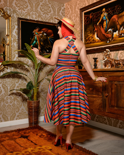 A woman stands indoors wearing a colorful striped halter dress with a flared skirt, a wide-brim straw hat, and a red headscarf. The room has patterned wallpaper, framed artwork, a potted plant, and wooden furniture.